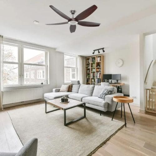 Salon lumineux avec canapé gris, table basse et bibliothèque; plafond équipé d’un ventilateur, ambiance chaleureuse et aérée. Destratificateur d'air? intégré par l’idée générale.