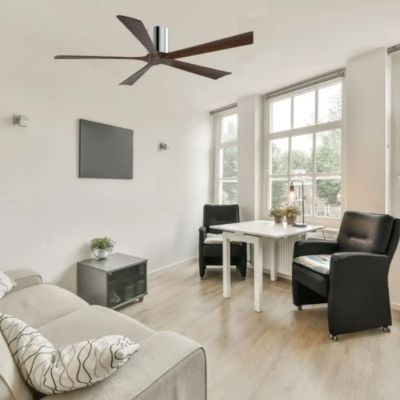 Salle de séjour lumineuse avec canapé, fauteuils et table près de grandes fenêtres et d'un ventilateur de plafond noir.