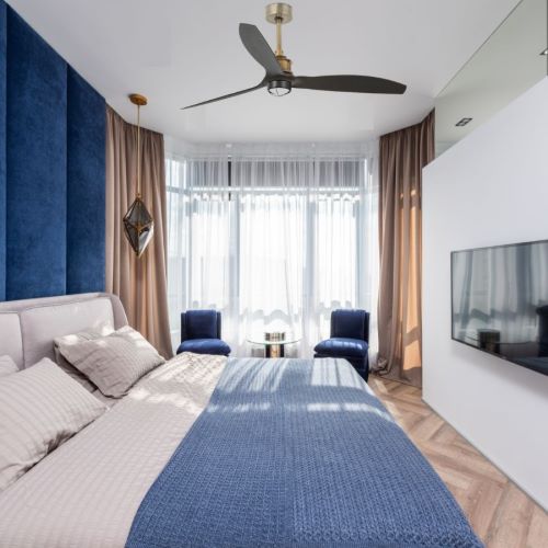 Ventilateur plafonnier noir à trois pales installé au-dessus d'un lit dans une chambre lumineuse.